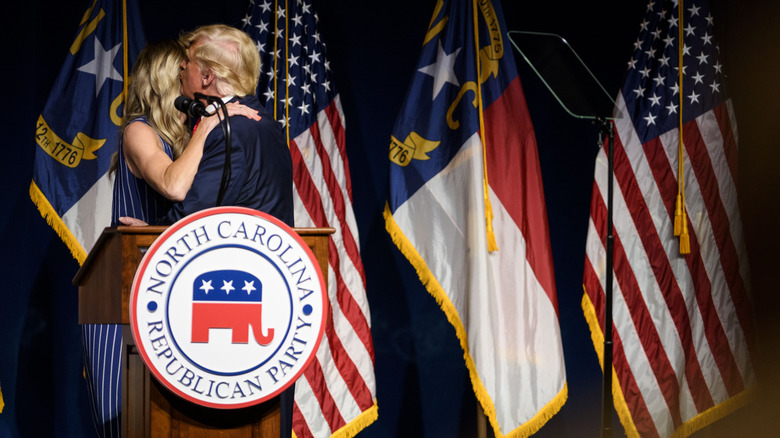 Donald Trump and Lara Trump holding each other close as he kisses her in front of flags at the 2021 North Carolina GOP Convention.