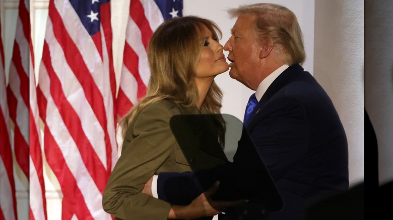 Melania Trump smiling and raising her chin as a sweaty Donald Trump opens his mouth to kiss her behind a teleprompter in the Rose Garden in 2020.