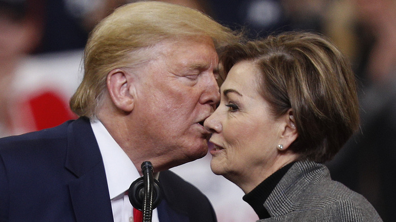 Kim Reynolds looking off to the side as Donald Trump's lips get close to her nose during a January 2020 rally.