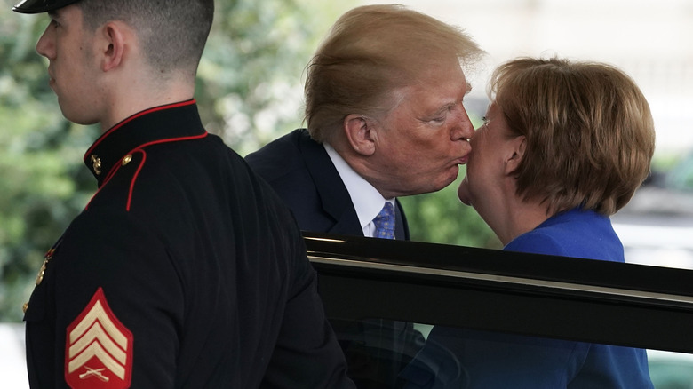 Donald Trump pushing his lips out far to kiss Angela Merkel beside a car in 2017.