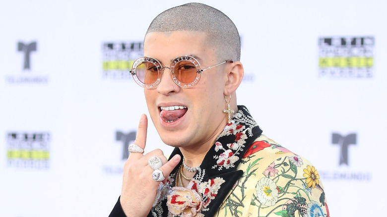 A younger Bad Bunny, with shaven head and sunglasses, appears in a multi-colored suit with a flower pattern and sticking his tongue out