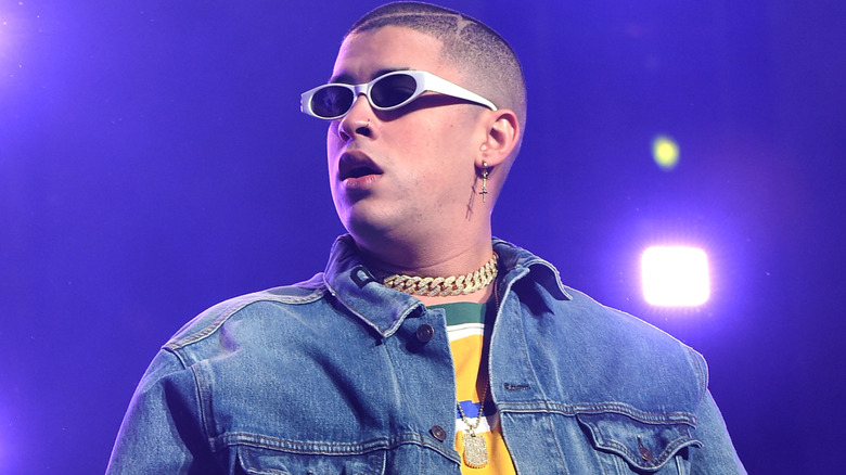 image of Bad Bunny wearing sunglasses, a buzz cut, a jacket, a thick necklace and a colorful shirt during a performance on stage