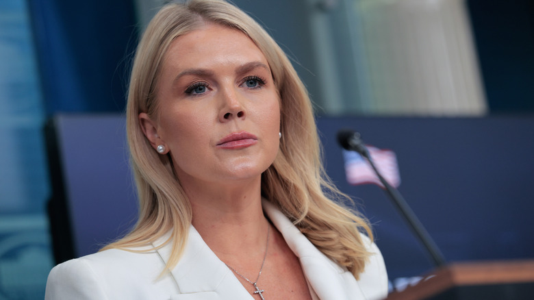 Karoline Leavitt looking serious behind the podium in a white blazer