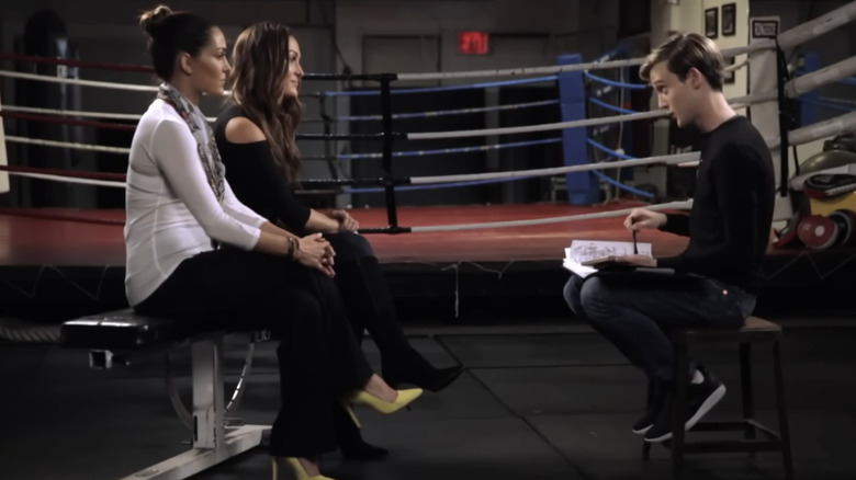Brie and Nicki Garcia sit in front of a wrestling ring facing Tyler Henry