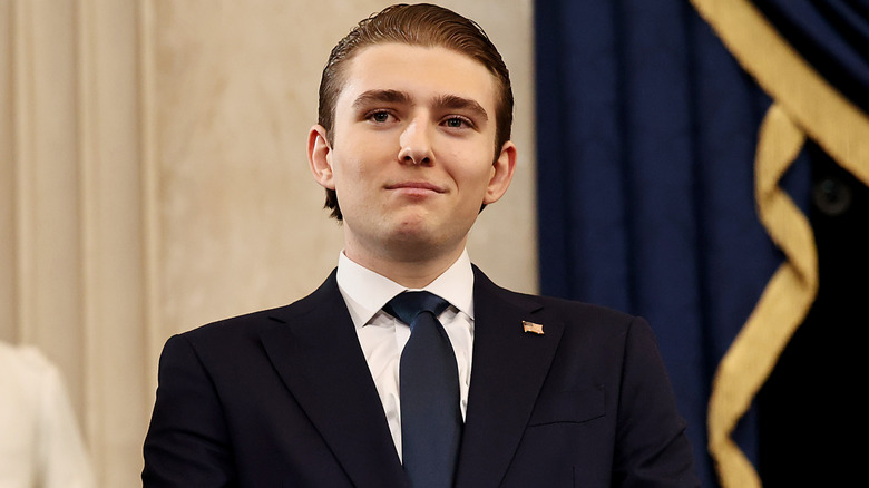 Barron Trump grinning in a black suit with a navy tie and his hair slicked back