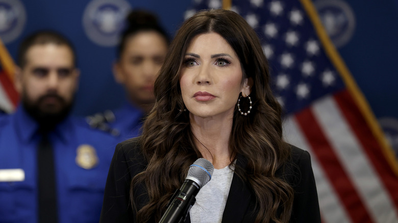 Kristi Noem standing at a microphone with police officers in background