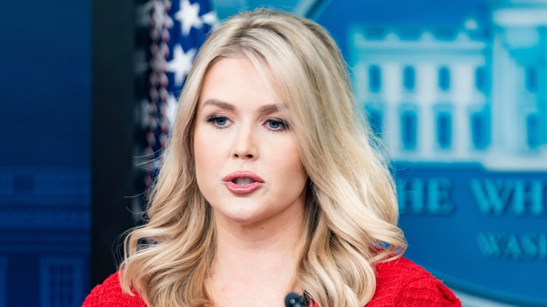 Karoline Leavitt speaking at a White House press briefing while wearing a red and black sweater.