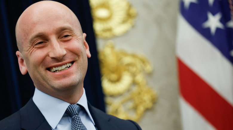 Stephen Miller grinning widely in a dark suit with an American flag behind him