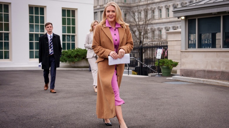 Karoline walking with a long brown trench coat and pink dress.