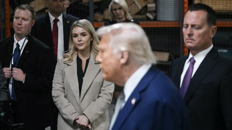 Karoline Leavitt smirking in a light tan trench coat while watching president Donald Trump speak at John F. Kennedy center.