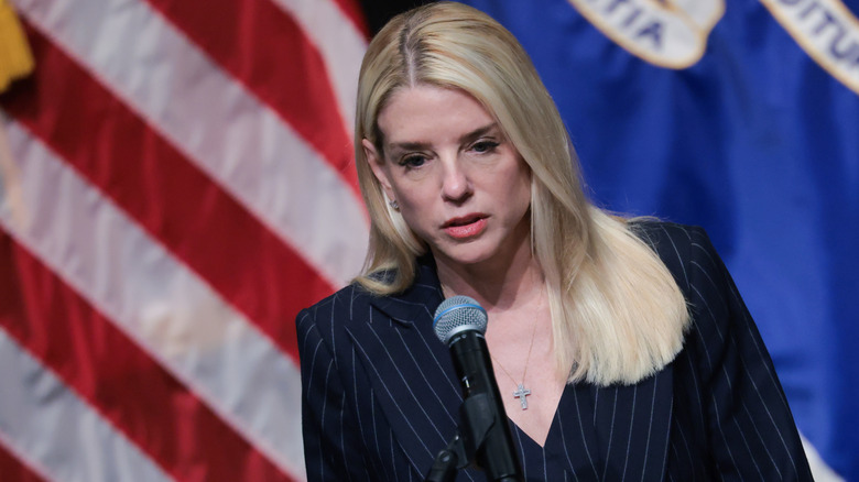 Pam Bondi looking confused in a pinstripe suit in front of a microphone