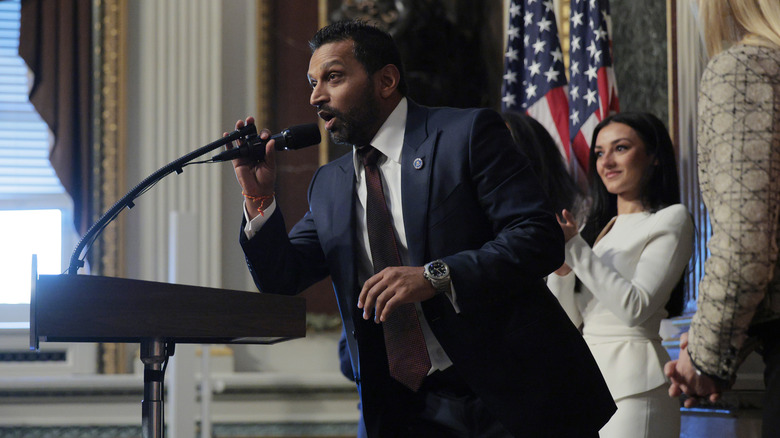 Kash Patel talks into a microphone with girlfriend, Alexis Wilkins standing behind him.