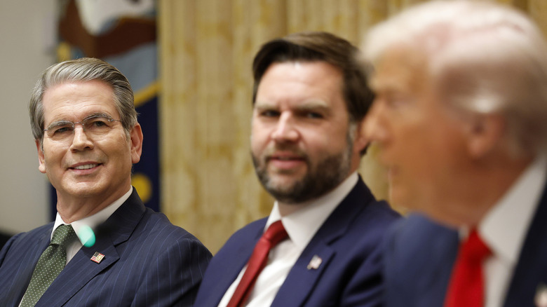 In focus, Scott Bessent next to JD Vance who looks over his shoulder in an unfocused image, looking toward Donald Trump