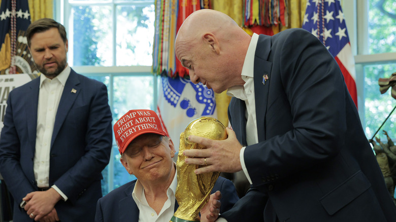 JD Vance, out of focus, looks down at Donald Trump, who is seated at his desk in the Oval Office as he is shown the FIFA World Cup.