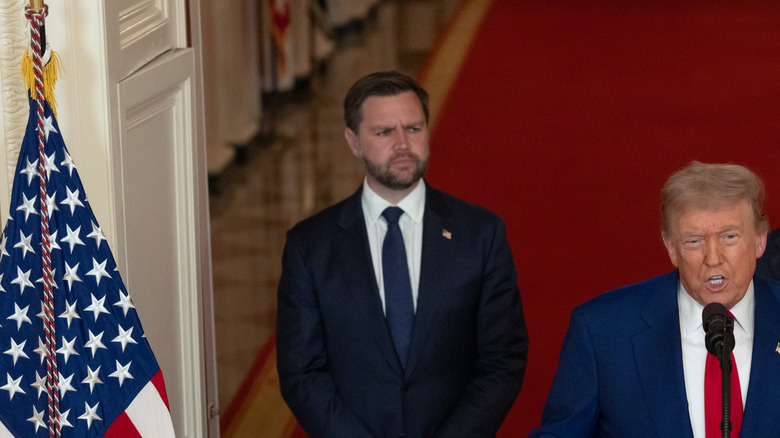 In the East Room hall of the White House, Donald Trump talks into a microphone with JD Vance standing behind him.