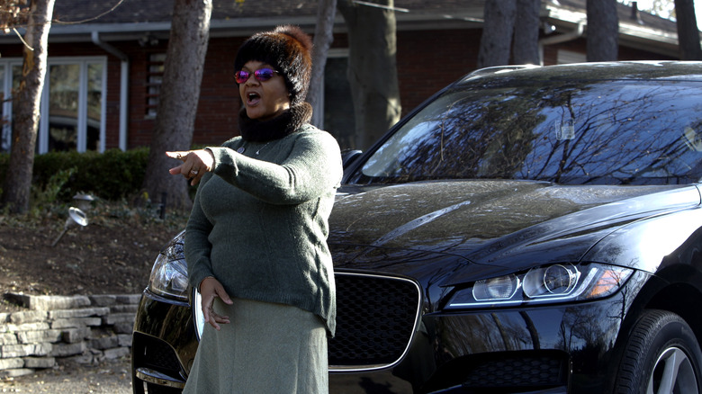 Monica Conyers in winter clothes, pointing with mouth open in front of car.