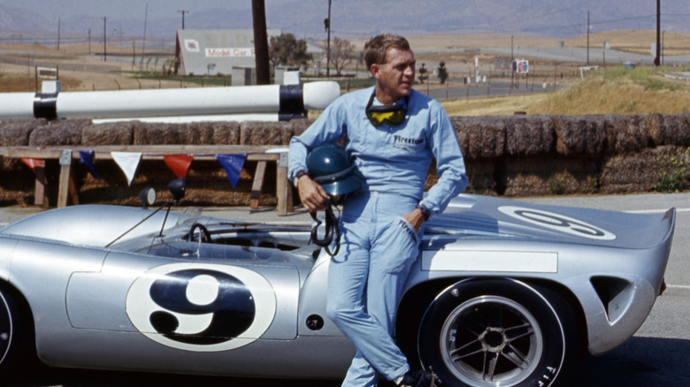 Steve McQueen wearing a racing stood and leaning against a racecar