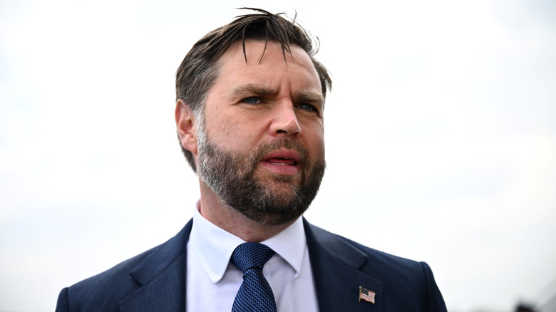 JD Vance before a cloudy white sky, while his hair flops onto his forehead, captured mid-talk in a suit.
