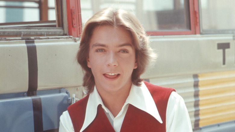 Young Cassidy with shoulder-length hair wearing a white shirt and red vest.