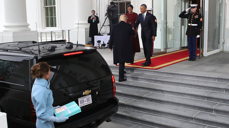 Melania Trump holding a gift beside a SUV while Donald Trump greets Barack and Michelle Obama at the White House.