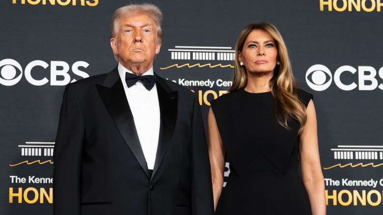 A stony-faced Donald Trump wears a tuxedo and poses beside Melania Trump, who is wearing a black dress.