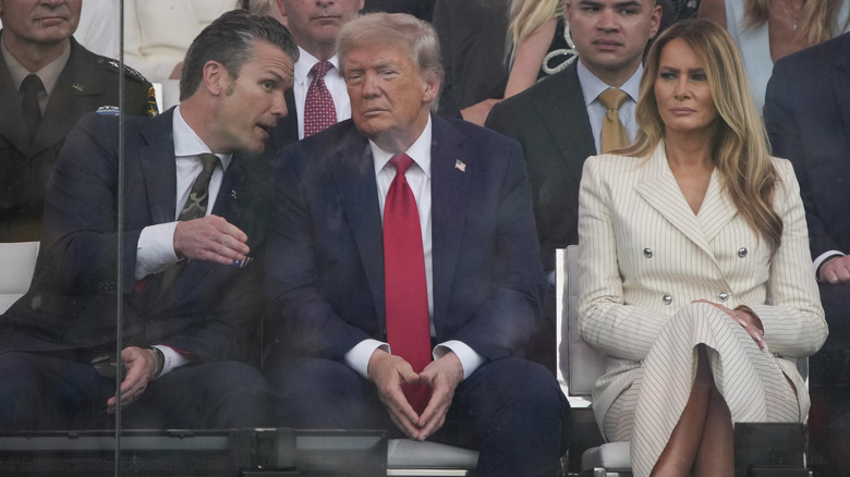 Pete Hegseth speaking to Donald Trump during the 2025 Army parade while Melania Trump frowns next to him.