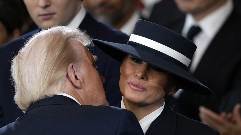 Donald Trump leaning in under Melania Trump's hat to give her an air kiss during his 2025 inauguration.