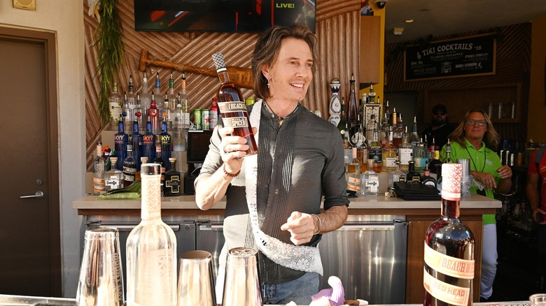 Rick Springfield standing behind a bar holding up a liquor bottle