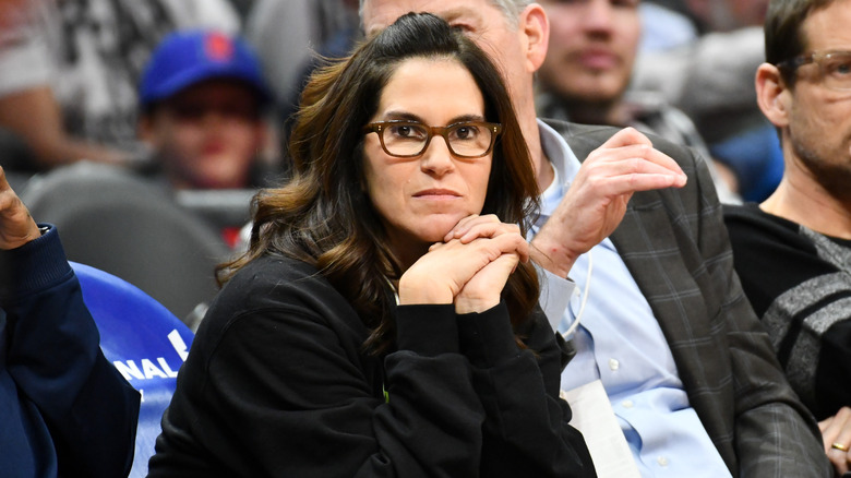 Jami Gertz sitting next to her husband, Tony Ressler, at an Atlanta Hawks game with her face on her hands with minimal makeup on
