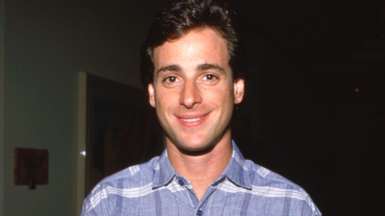 Bob Saget smiling at a 1990 event, wearing a blue checked shirt