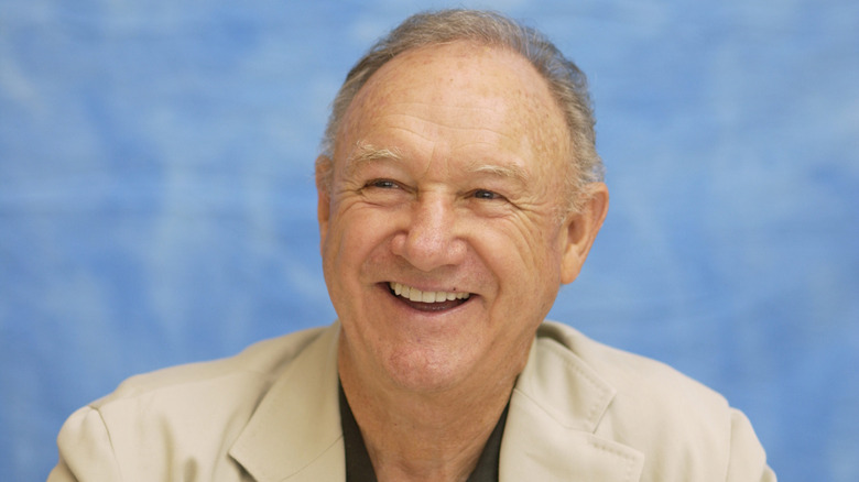 Gene Hackman smiling in a tan blazer at a press conference