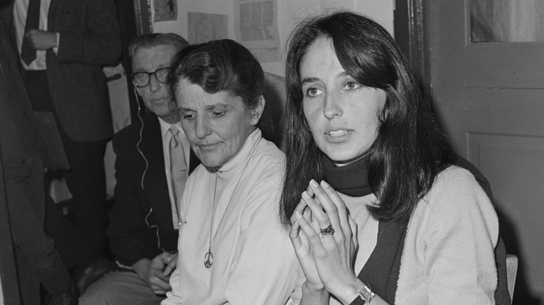Black and white photo of Joan Baez wearing a scarf with her hands folded sitting beside her mother in a white shirt and peace necklace