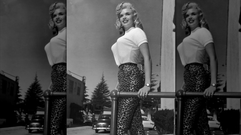 Jayne Mansfield in white sweater and leopard pants holding onto a rail and smiling