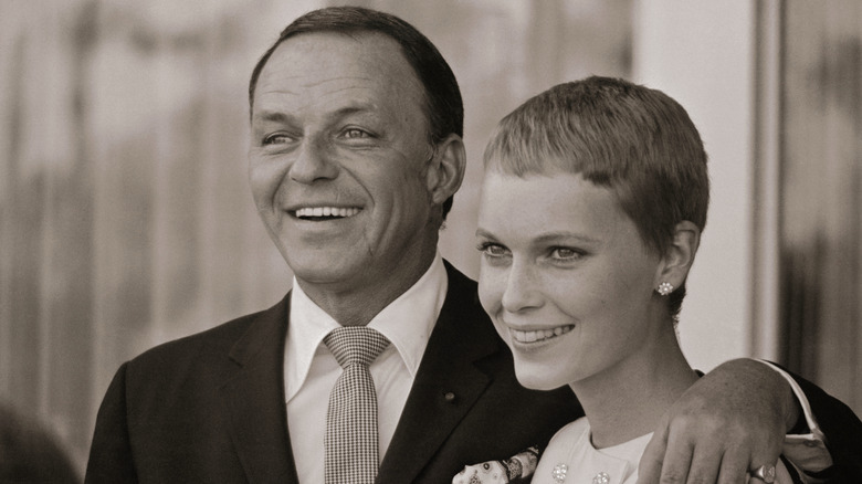 Black and white photo of Frank Sinatra in a suit and tie with his arm around Mia Farrow with a pixie cut both smiling