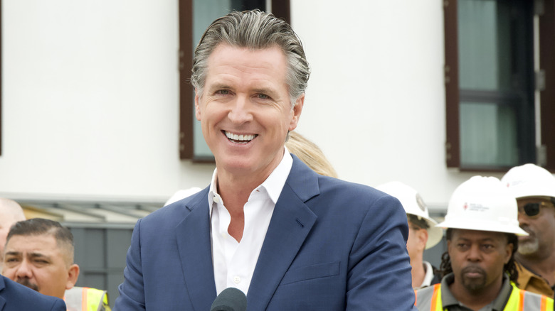 Gavin Newsom laughing in front of a mic in a dark grey suit jacket and a white shirt