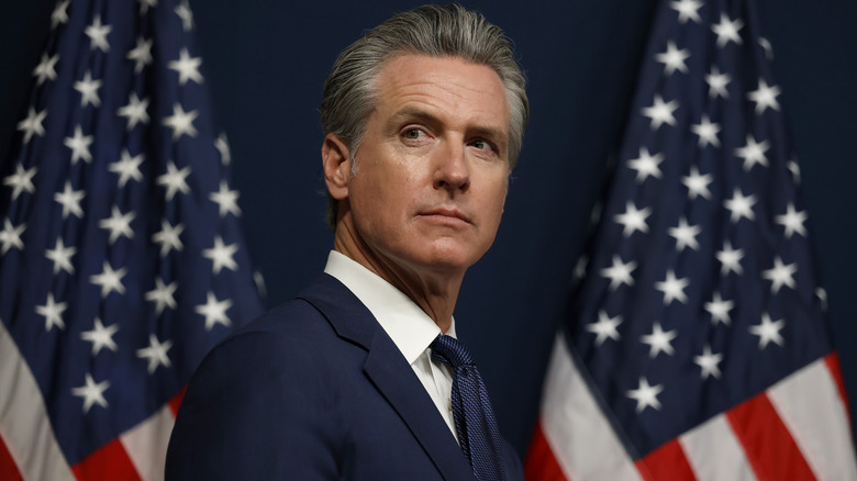 Gavin Newsom looking pensive in a navy suit in front of two American flags