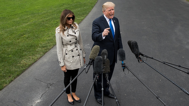 Melania Trump wears a light tan trench coat and sunglasses while standing beside Donald Trump, who is speaking into a bunch of microphones.