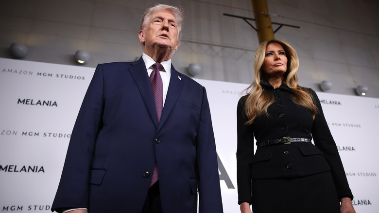 Donald Trump wears a dark blue suit and Melania Trump wears a black suit dress as they both stand with their arms straight at their side at the "Melania" documentary premiere.