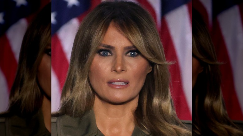 Melania Trump wearing dark eye makeup, shiny lip gloss, and heavy bronzer with a deer-in-the-headlights expression.