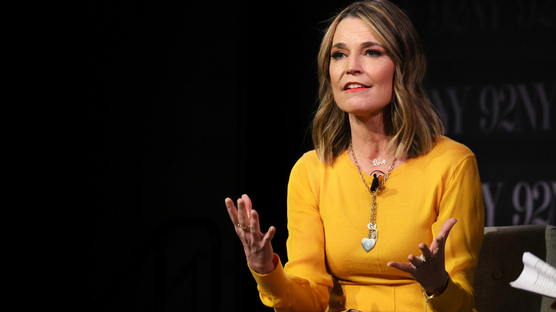 Savannah Guthrie speaking at an event while wearing a yellow dress and heart pendant necklace.