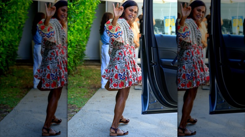 Mindy Kaling smiling and waving next to an open car door