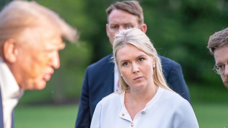 Karoline Leavitt outside, head tilted, staring at out-of-focus Donald Trump as he speaks