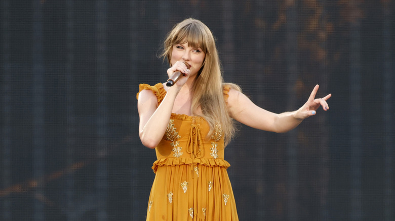 Taylor Swift wearing an orange dress and singing on stage, wearing her hair styled down with bangs.