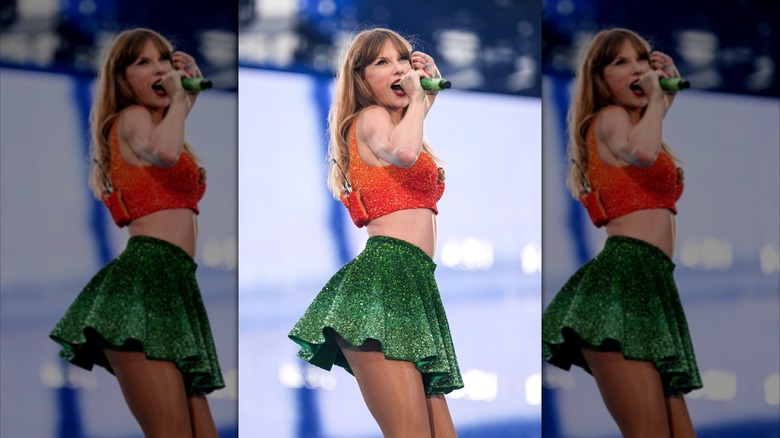 Taylor Swift singing on stage holding a microphone, hair down bangs, orange bedazzled crop top and green bedazzled skirt