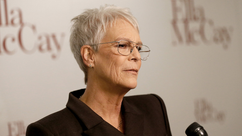 Jamie Lee Curtis wearing glasses and a dark suit in front of a microphone