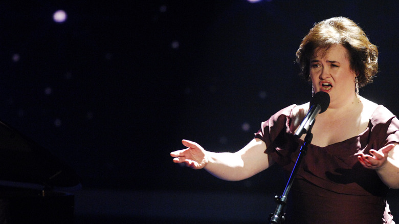 Susan Boyle holding her arms up while performing onstage in a dark purple dress