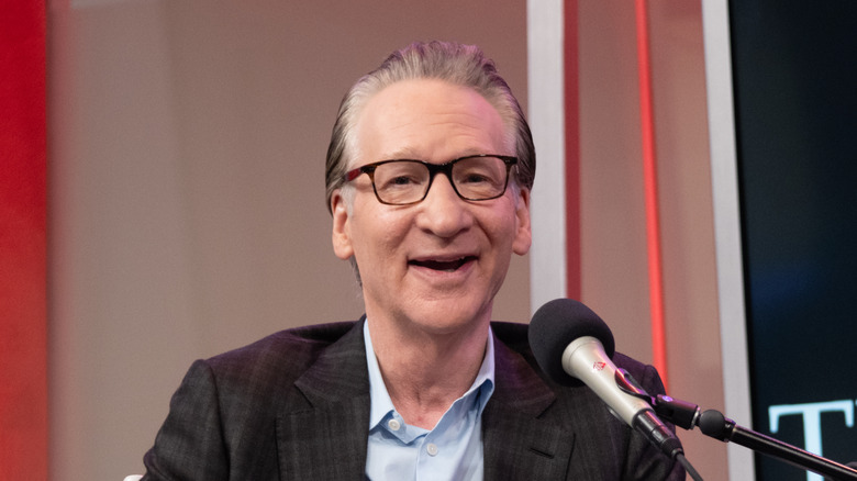 Bill Maher in a checked coat before a mic