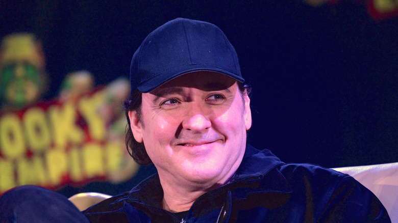 John Cusack smiling in a cap and jacket