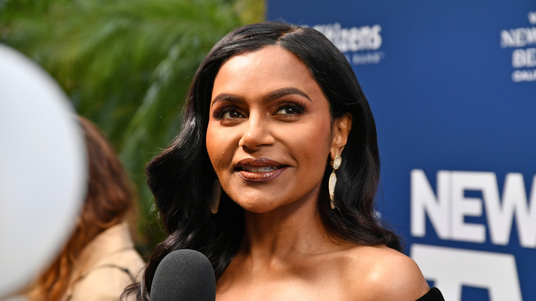 Mindy Kaling speaking into a mic in off-shoulder outfit