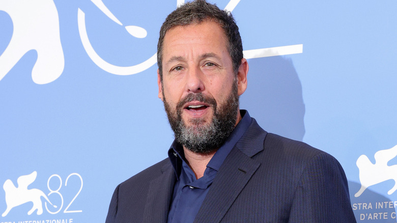 Adam Sandler posing for a photo in a dark suit with no tie